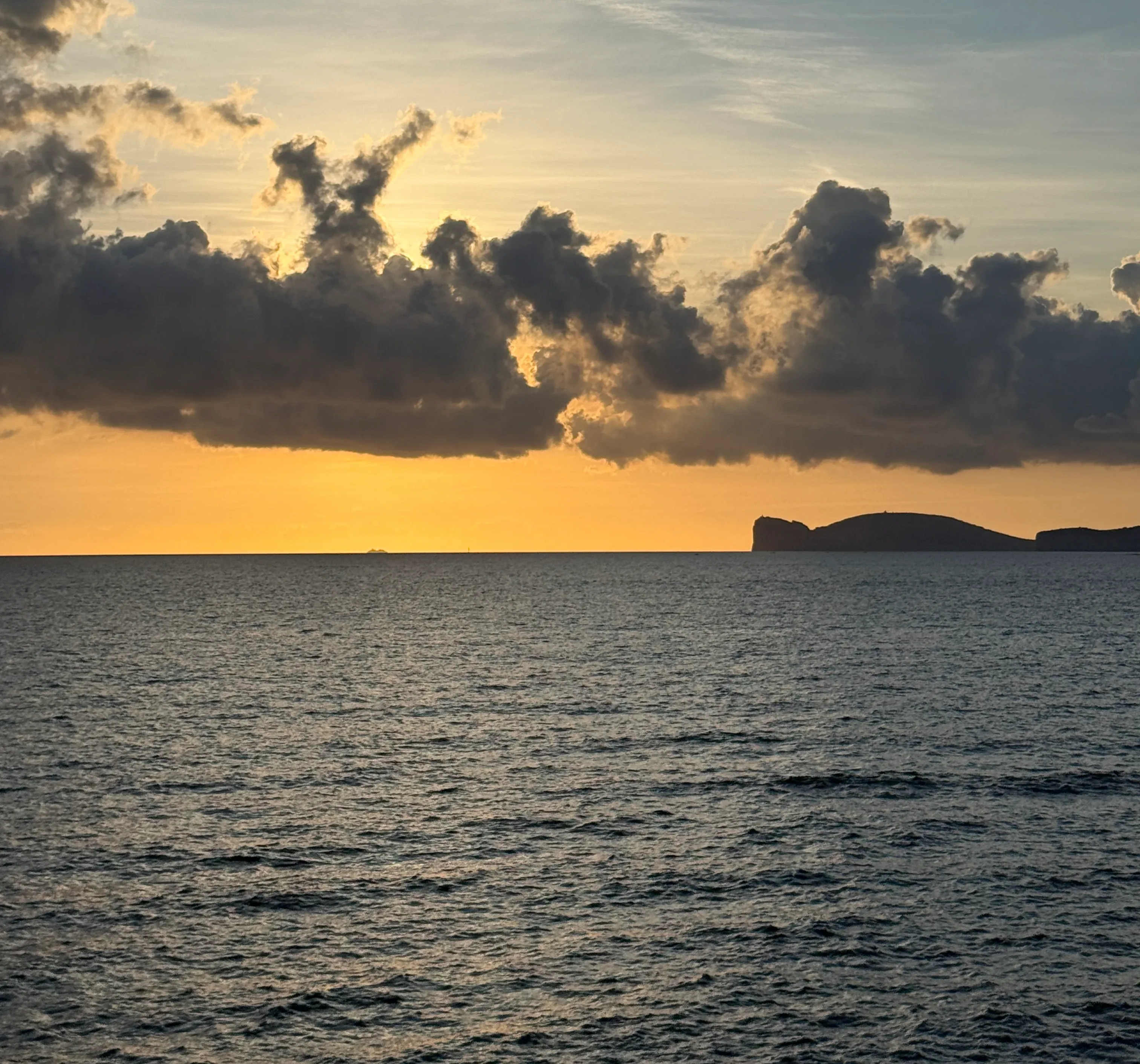 Dark, backlit clouds over a golden horizon and calm sea, with a headland in silhouette.
