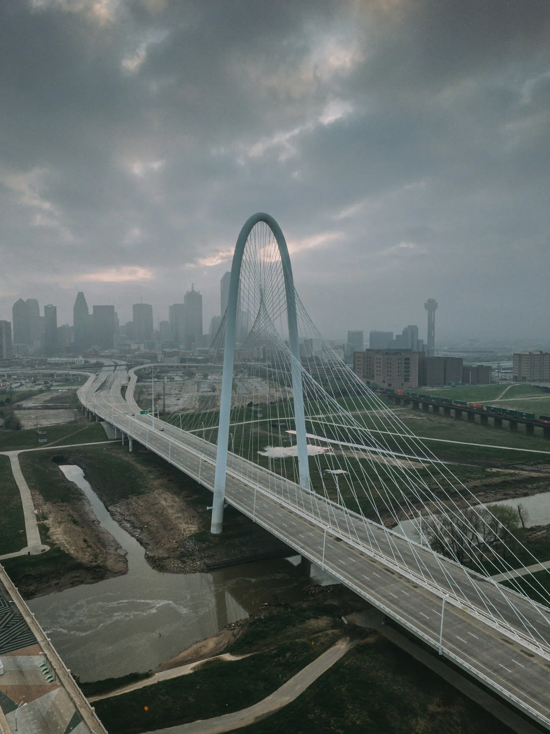 A city skyline with a modern cable-stayed bridge in the foreground, featuring a large arch and multiple cables, under a cloudy sky.