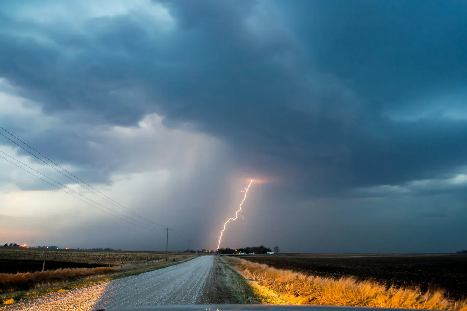 A rural gravel road under a dark stormy sky with a lightning bolt in the distance.